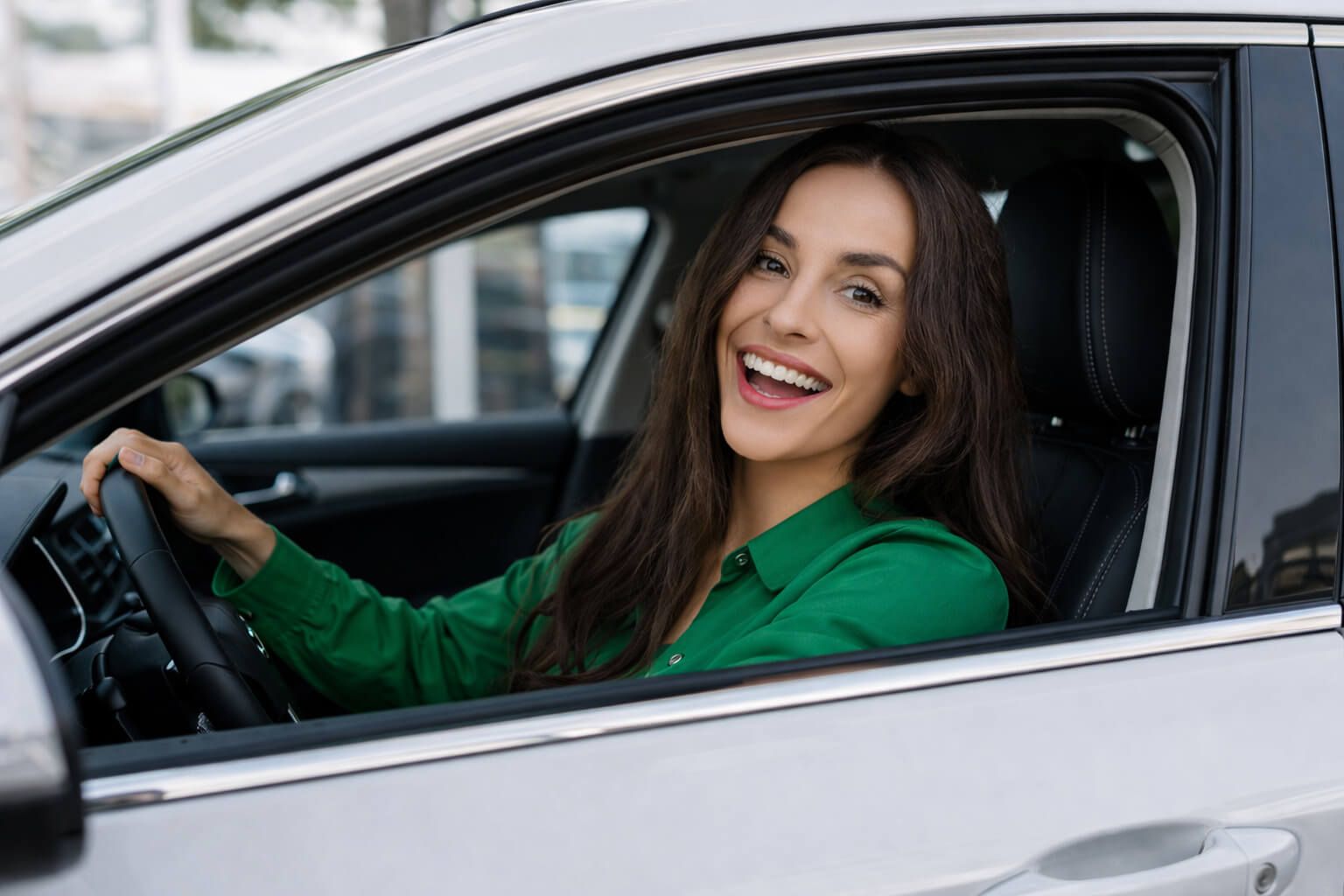 Smaidīga sieviete sēž pie automašīnas stūres ar atvērtu logu pilsētas vidē.