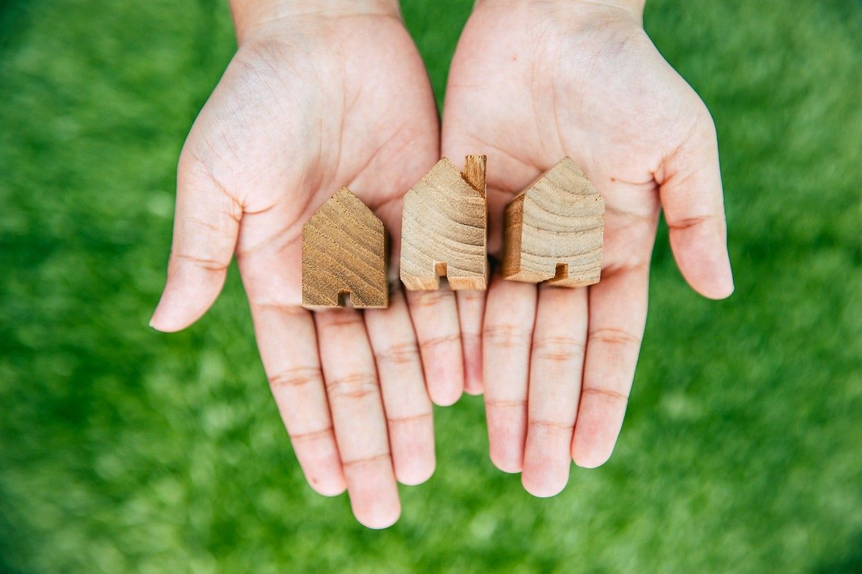 Две руки, держащие три маленьких деревянных домика, отражает сравнение кредитов и выбор наиболее подходящего предложения.