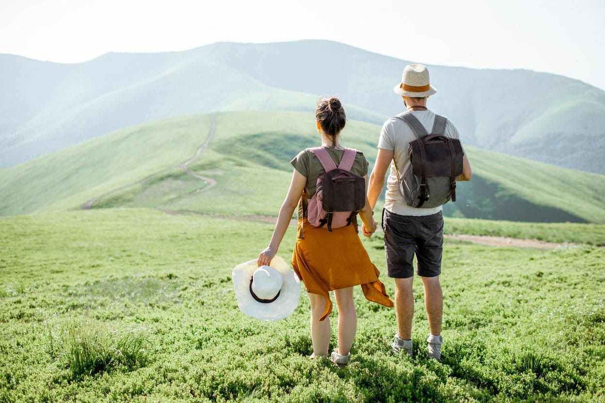 Пара с рюкзаками и шляпой в руке любуется зелёными горами, наслаждаясь спокойным осенним походом на природе.